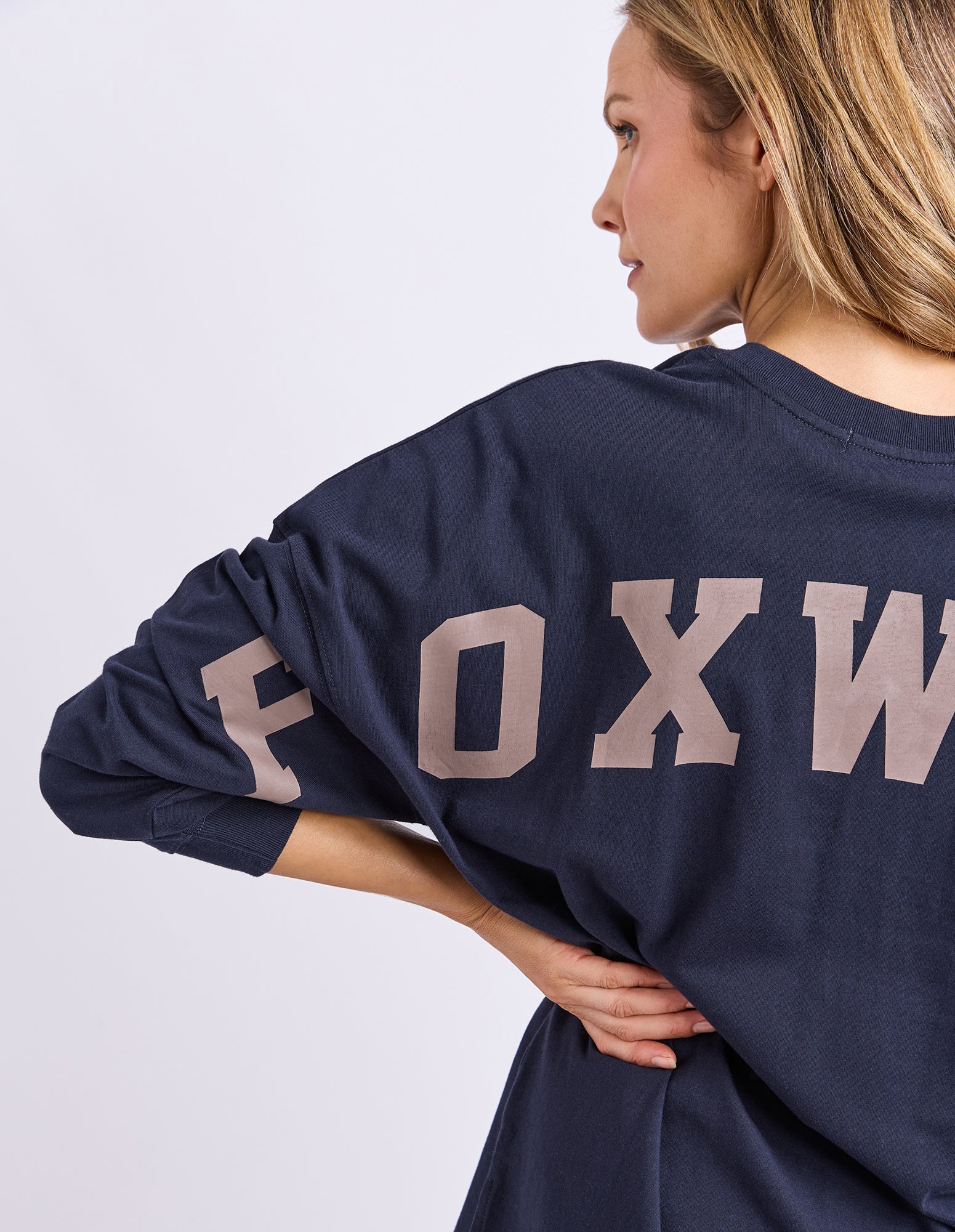 Image of Woman wearing a navy blue long sleeve tee with 'FOXWOOD' printed on the back in brown against a light grey background