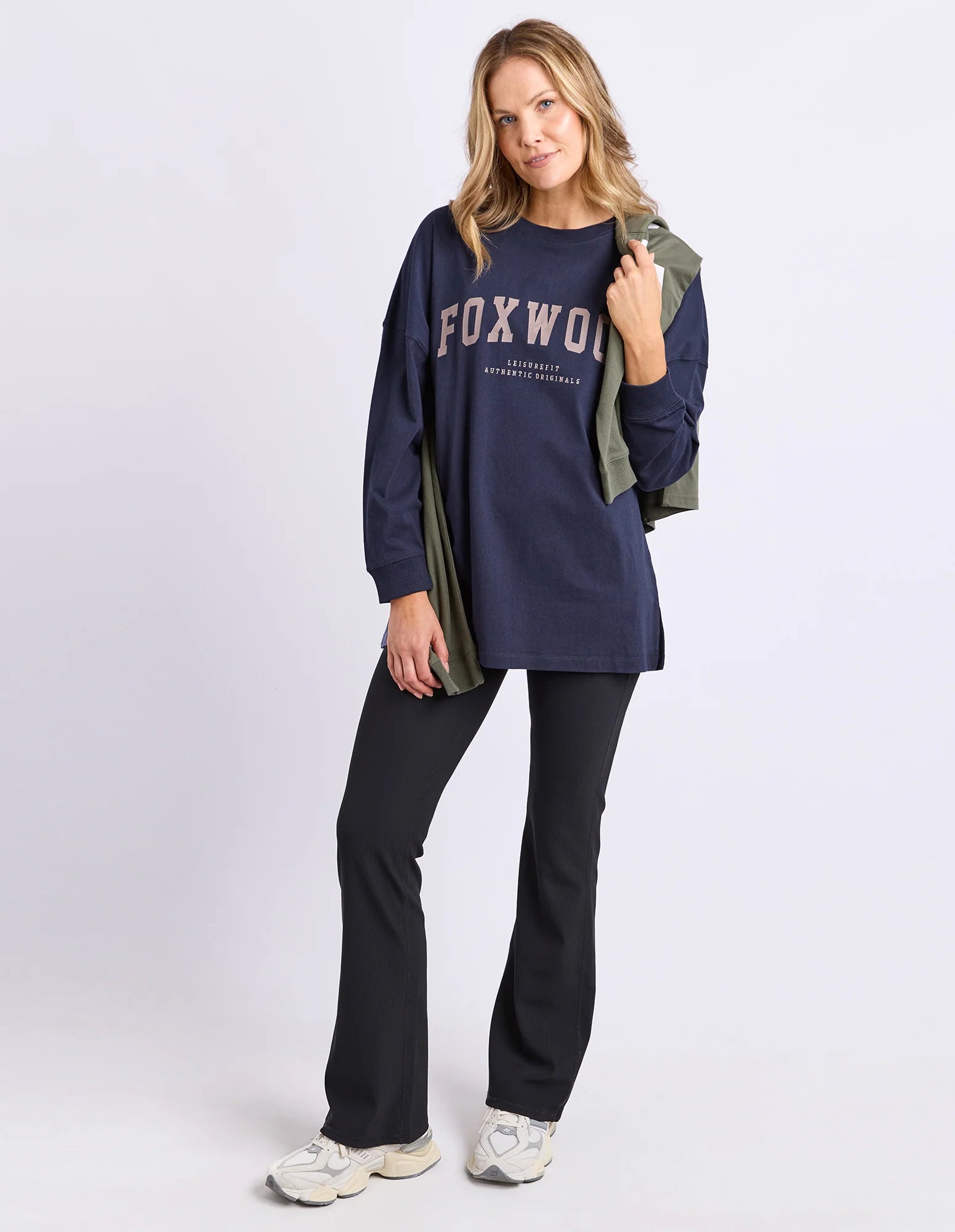 Woman wearing a navy long sleeve tee with 'FOXWOOD' branding in brown , black pants, and white sneakers on a light grey background.