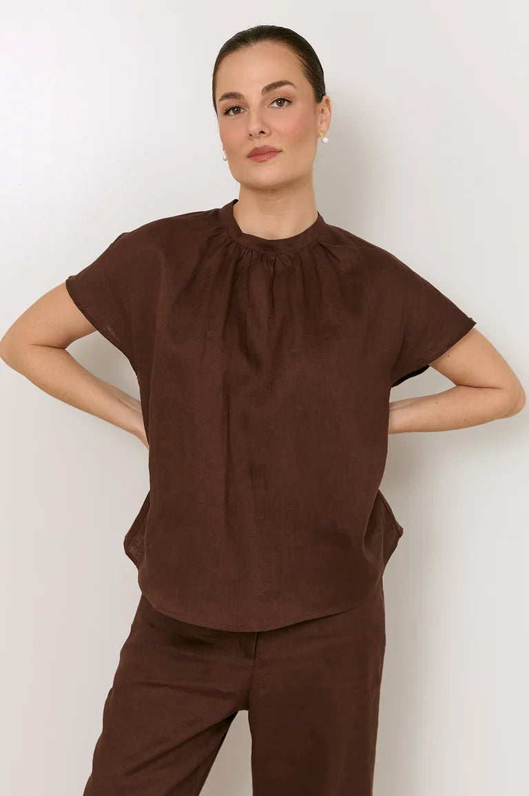 A woman modeling a Adorne dark brown short-sleeve top with a gathered, pleated neckline, paired with matching brown pants, against a plain light gray background.