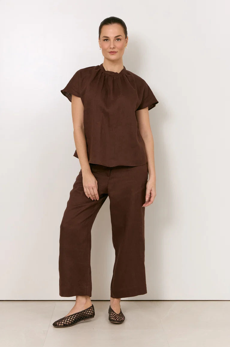 A woman modeling a Adorne matching brown two-piece outfit consisting of a short-sleeve top with a gathered neckline and wide-leg cropped pants, paired with black patterned flats, against a plain white studio background.