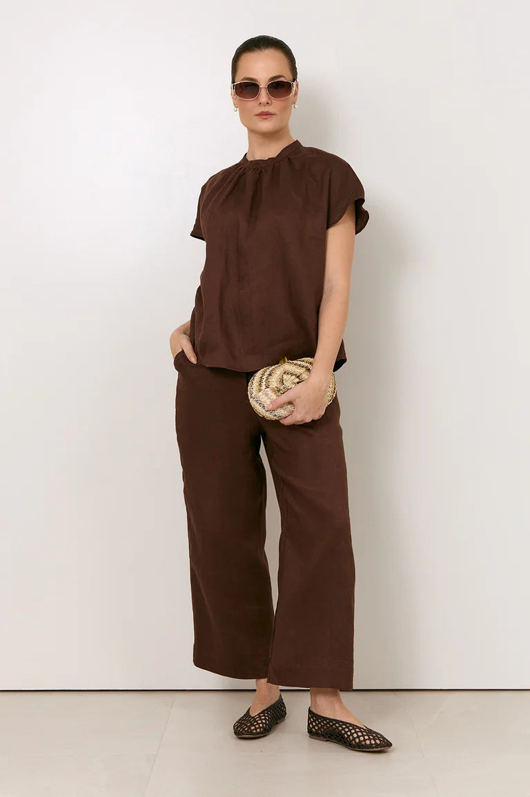 Image of A woman modeling a Adorne matching brown two-piece outfit consisting of a short-sleeve top with a gathered neckline and wide-leg cropped pants, styled with sunglasses, a woven clutch, and flat shoes against a plain white wall.