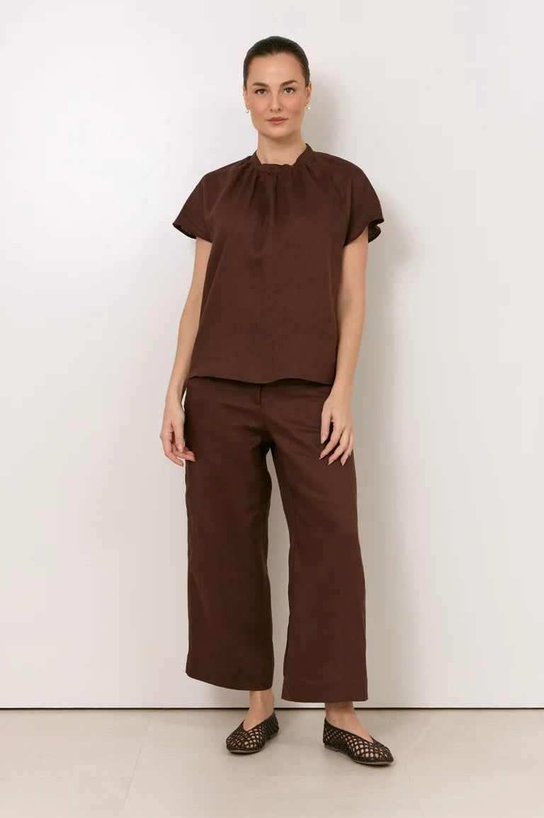 A woman modeling a Adorne matching brown two-piece outfit consisting of a short-sleeve top with a gathered neckline and wide-leg cropped pants, paired with brown woven flats, against a plain white studio background.