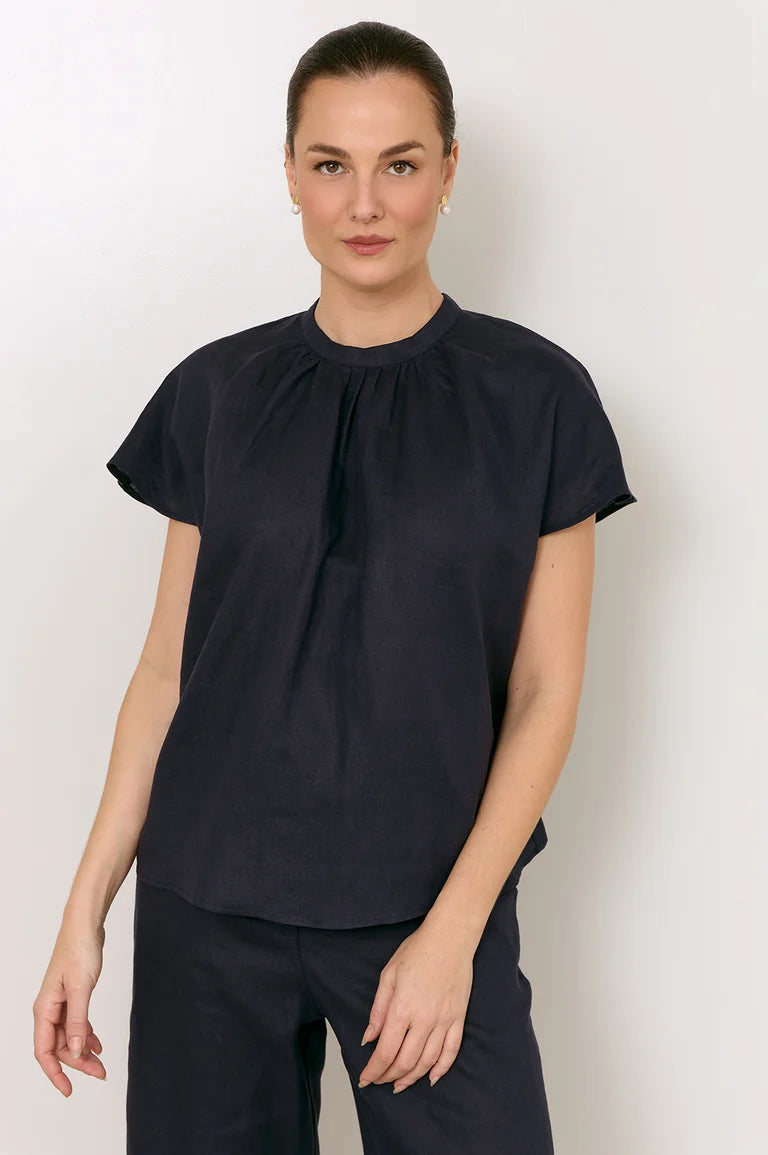 A woman modeling a Adorne short-sleeve black blouse with a gathered, pleated front neckline, paired with black trousers, against a plain light gray studio background.
