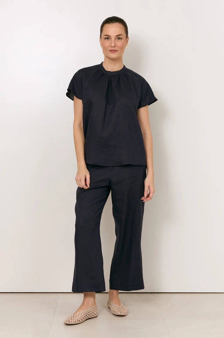 A woman stands against a plain off-white wall wearing a Adorne dark navy short-sleeve top with a gathered neckline and matching dark navy wide-leg cropped pants, paired with beige flat shoes.