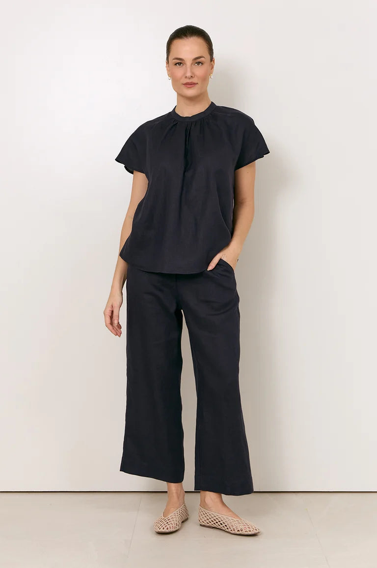 A woman modeling a Adorne dark navy two-piece set consisting of a short-sleeve blouse with a gathered neckline and matching wide-leg cropped pants, paired with beige woven flat shoes against a plain off-white studio background.