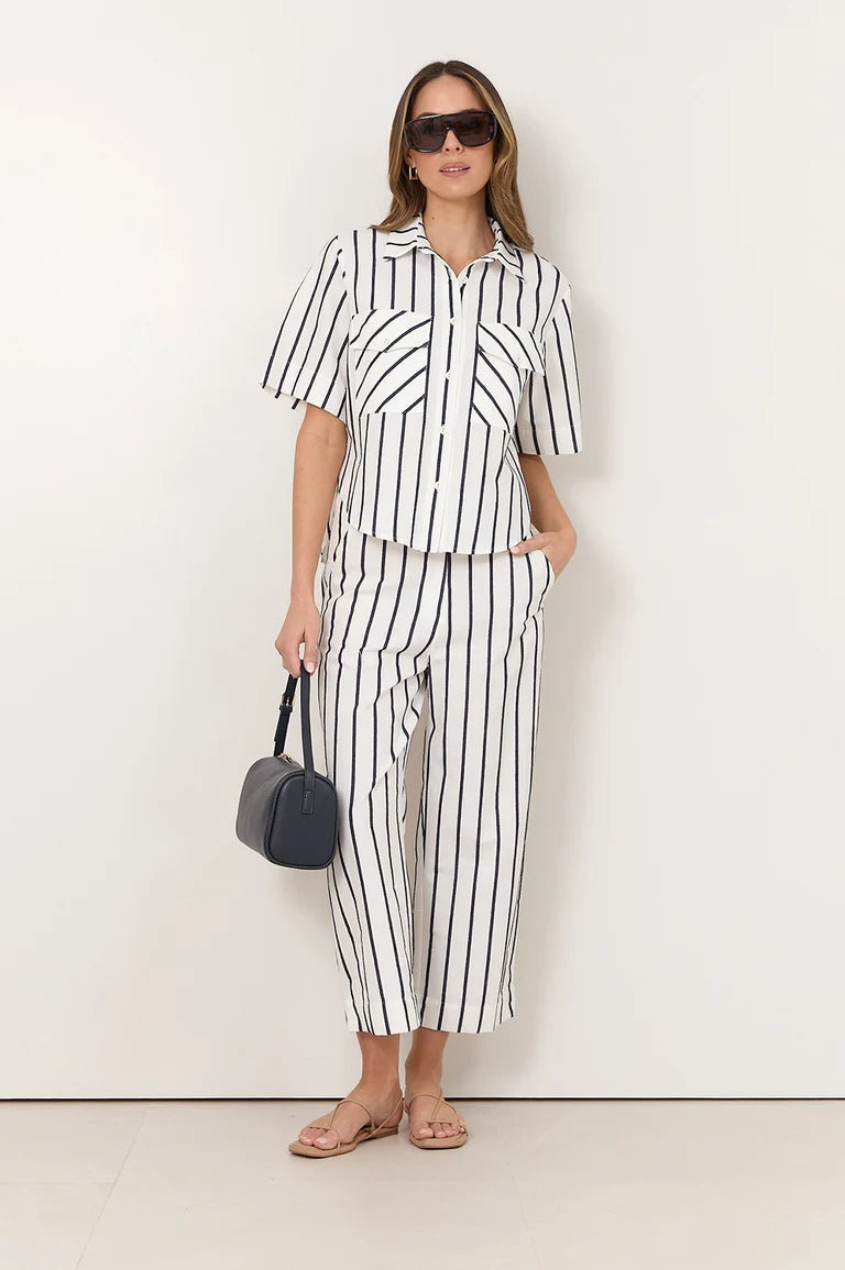 Image of A woman modeling a Adorne matching two-piece set with vertical black stripes on a white background, featuring a short-sleeve button-up shirt and wide-leg cropped pants. She is wearing black sunglasses, holding a small black handbag, and standing against a plain off-white wall.