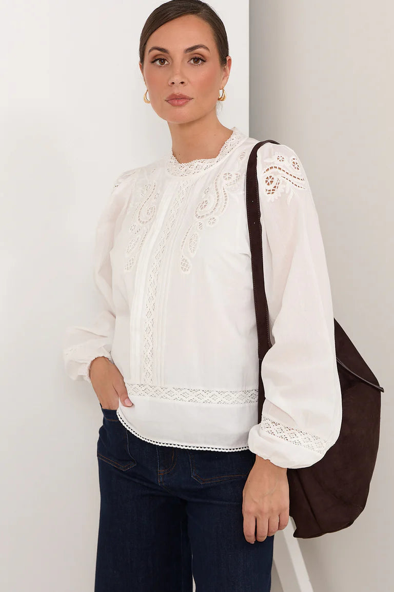 Image of Woman wearing a Adorne white blouse with lace details and dark jeans, holding a brown bag against a plain background.