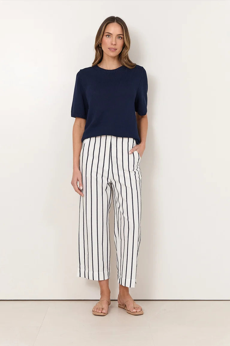 A woman modeling a Adorne navy blue short-sleeve top and white cropped pants with thin black vertical stripes, standing against a plain off-white wall.