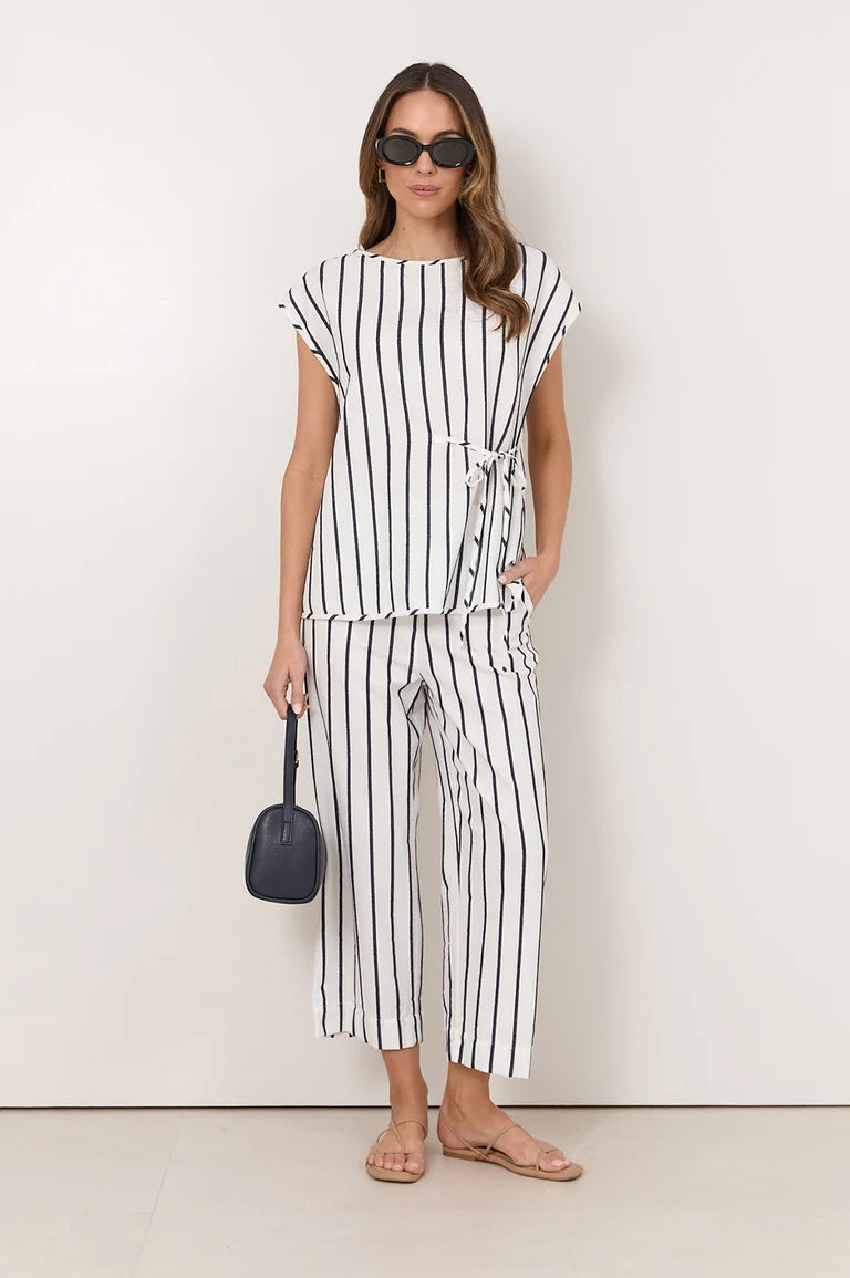 A woman modeling a Adrone matching two-piece set with vertical black stripes on a white base, featuring a short-sleeve top with a tie detail at the waist and matching cropped pants. She is wearing black sunglasses, holding a small black handbag, and standing against a plain off-white studio background.