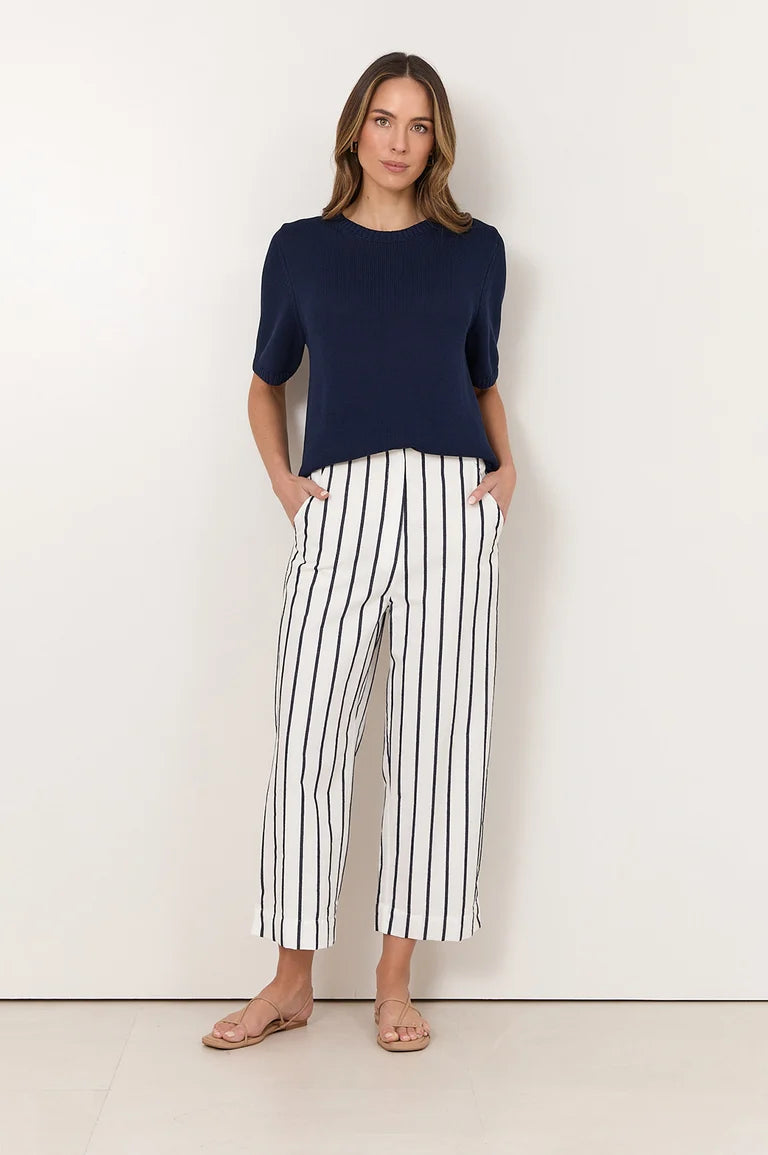 A woman modeling a navy blue short-sleeve top and Adorne white cropped pants with thin black vertical stripes, standing against a plain off-white wall.