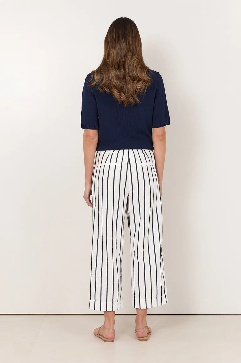 Image of A woman viewed from behind wearing a navy blue short-sleeve top and Adorne white cropped pants with thin black vertical stripes, standing against a plain off-white wall.