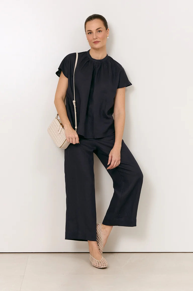 Image of A woman models a Adorne black short-sleeve top and matching wide-leg cropped pants, styled with a cream woven shoulder bag and beige studded flats against a plain white studio wall.