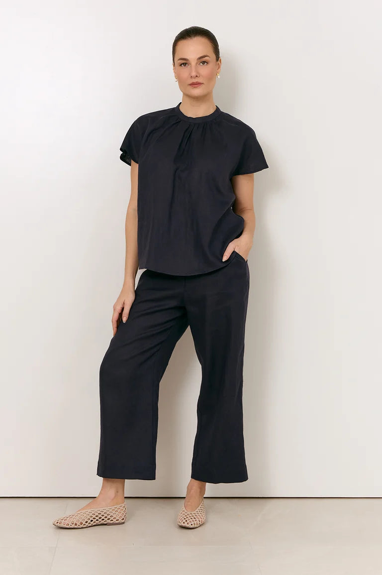 Image of A woman modeling a Adorne navy short-sleeve blouse with a gathered neckline and matching navy wide-leg cropped pants, paired with beige woven flat shoes, against a plain off-white studio background.
