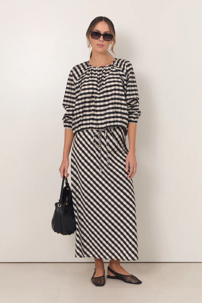 Image of A woman modeling a Adorne black and white striped two-piece set consisting of a long-sleeve top with a gathered neckline and a matching midi skirt with diagonal stripes. She is wearing black sunglasses, a black handbag, and black flats against a plain off-white studio background.