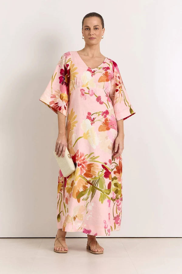 Image of A woman modeling a Adorne light pink kaftan dress with a vibrant floral print in red, orange, and green, holding a small cream clutch against a plain white studio background.