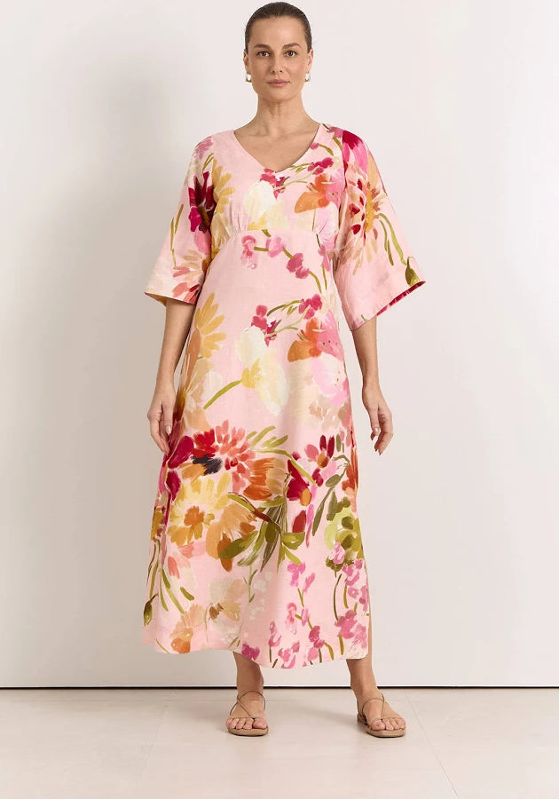 Image of A woman modeling a Adorne pink kaftan dress with a vibrant watercolor floral print in red, orange, yellow, and green, set against a plain light gray studio background.