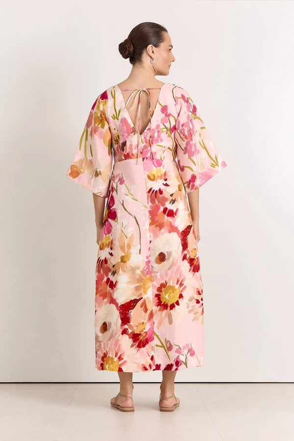 Image of A woman shown from the back wearing a Adorne light pink kaftan-style dress with a large watercolor floral print in red, orange, and cream, featuring wide short sleeves and a tie-back neckline.