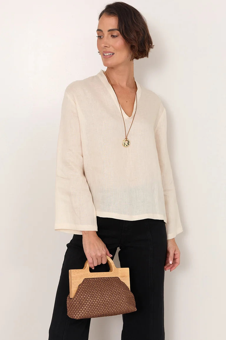 A woman wearing a cream-colored, long-sleeved linen blouse and black pants, holding a Adorne brown and beige woven handbag against a plain white background.