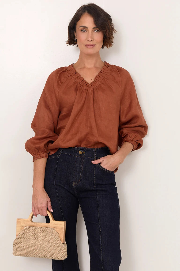 Image of A woman modeling a rust-brown long-sleeve blouse with a gathered V-neck, paired with dark blue jeans and holding a Adorne woven straw handbag against a plain off-white background.
