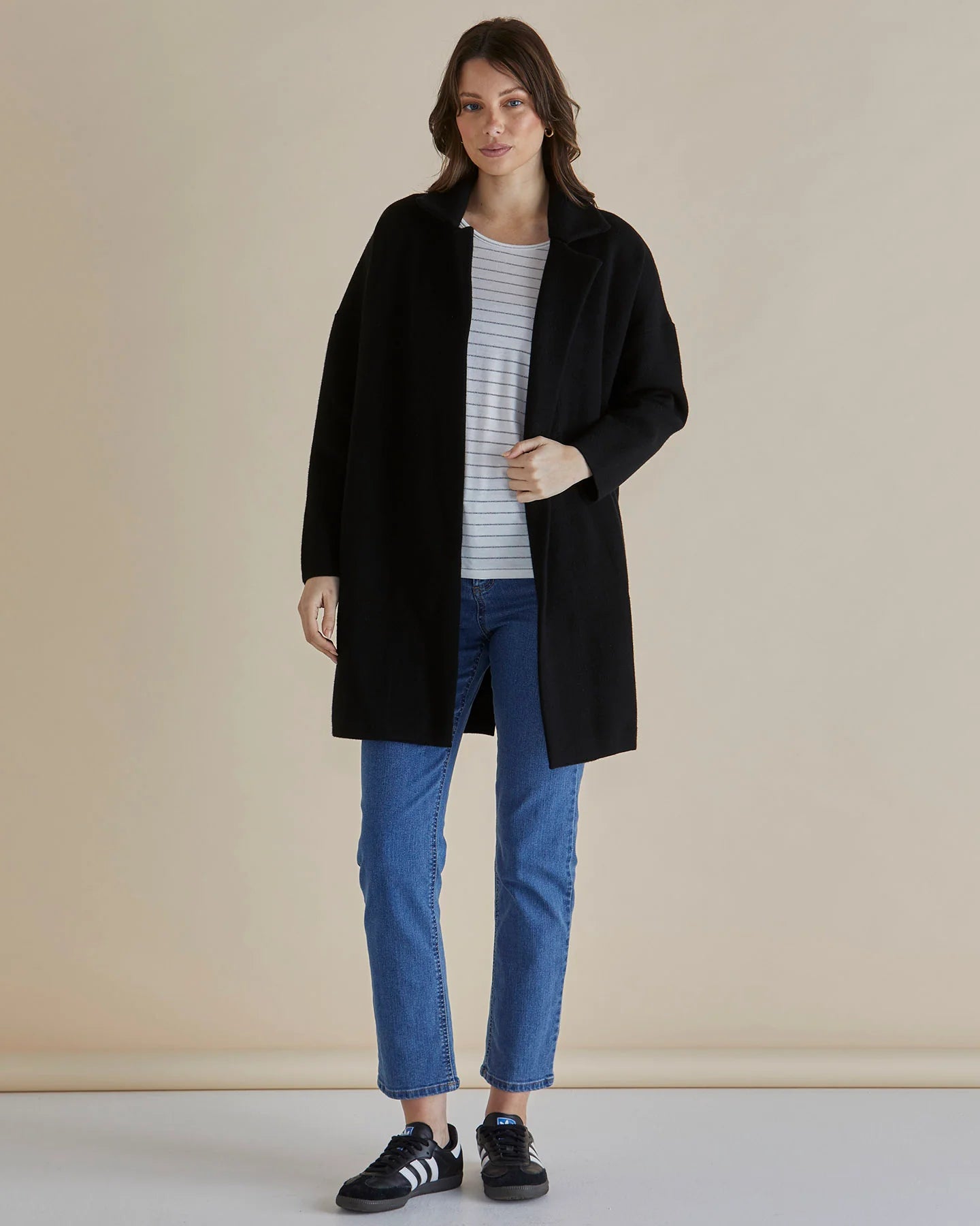 Image of Woman wearing a Betty Basics black coat, striped shirt, and blue jeans on a beige background