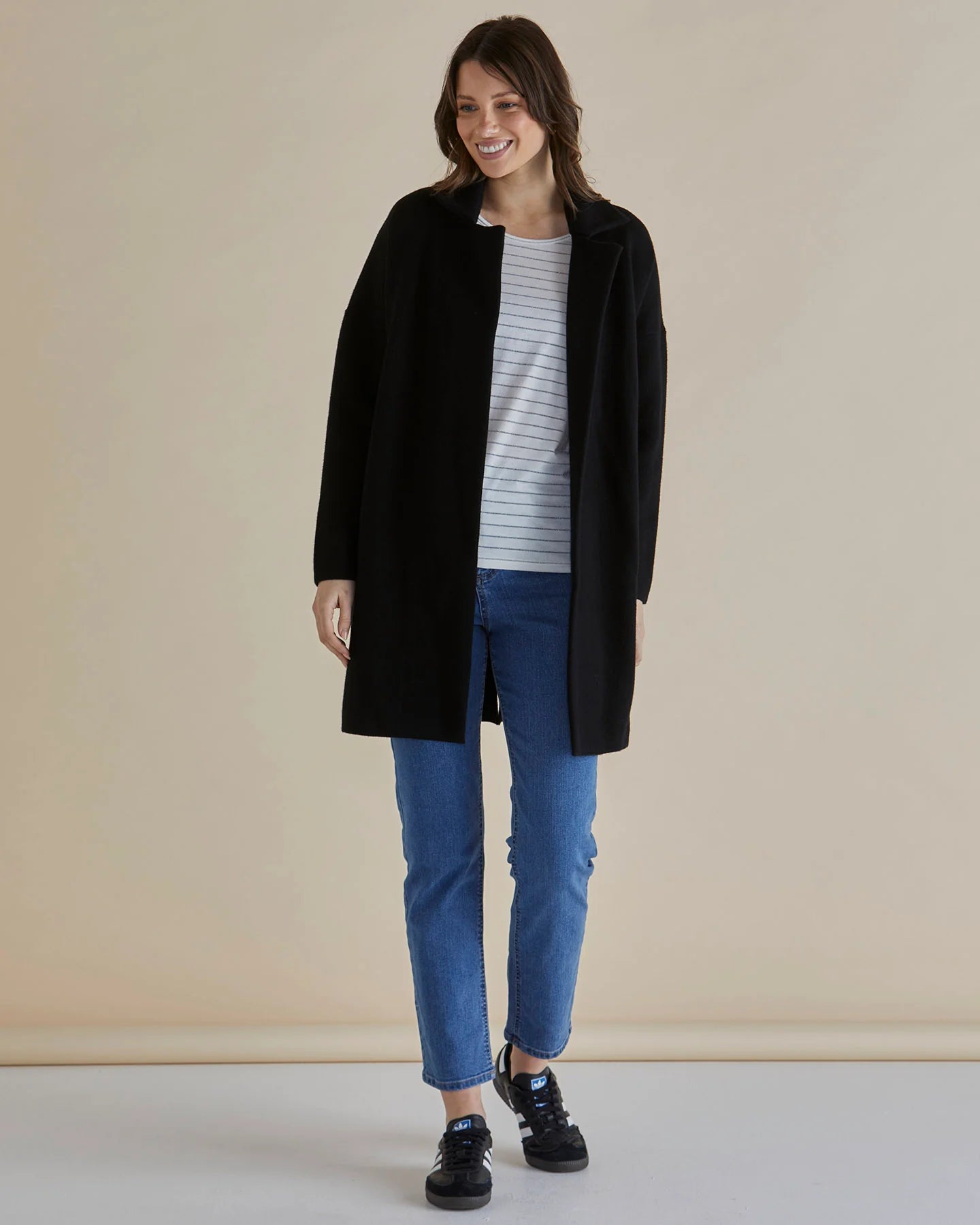 Image of Woman wearing a Betty Basics black coat, gray striped shirt, and blue jeans against a beige background