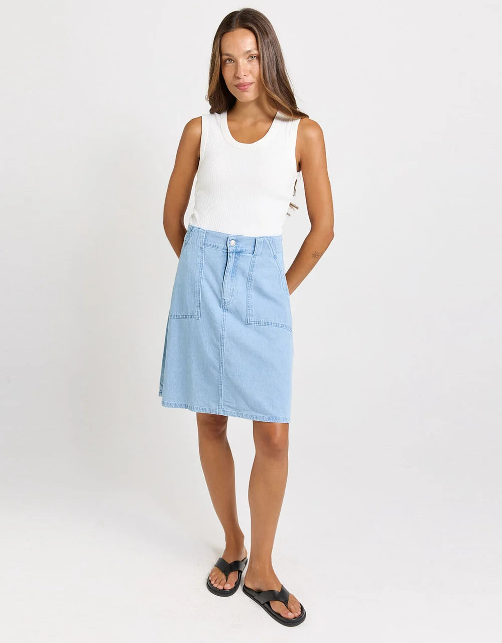 A woman modeling a Betty Basics light blue denim A-line skirt with a white sleeveless top and black sandals, standing against a plain white studio background.