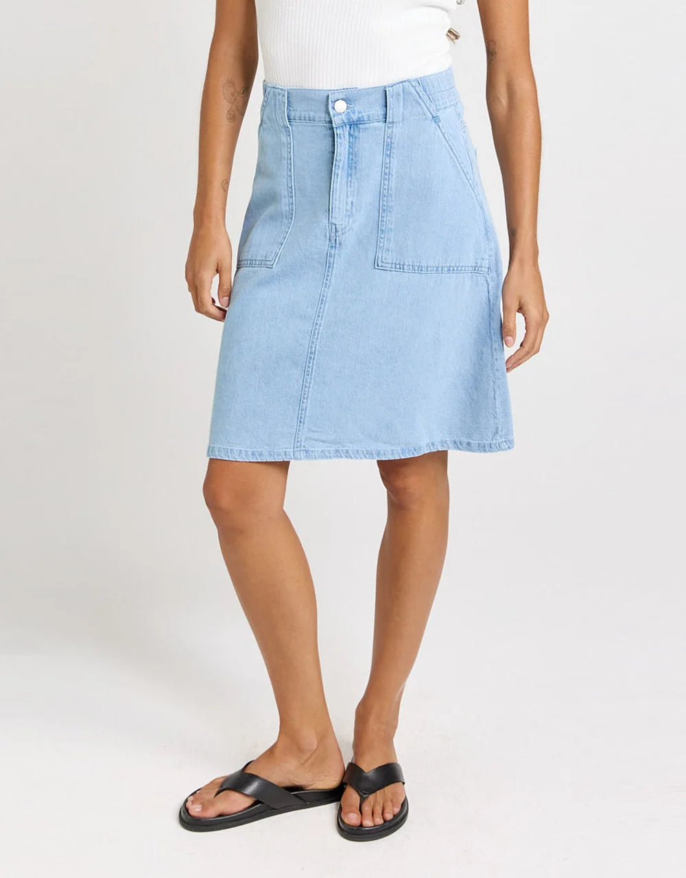 A model wearing a Betty Basics light blue denim A-line skirt with front patch pockets, paired with a white ribbed top and black slide sandals, against a plain white studio background.