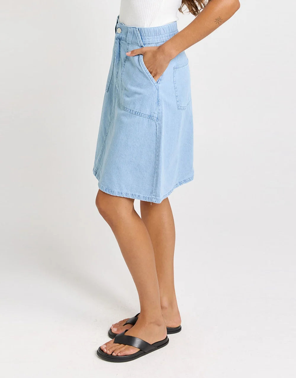 Image of A model wearing a Betty Basics light blue denim A-line skirt with front patch pockets, styled with a white ribbed top and black slide sandals against a plain white studio background.