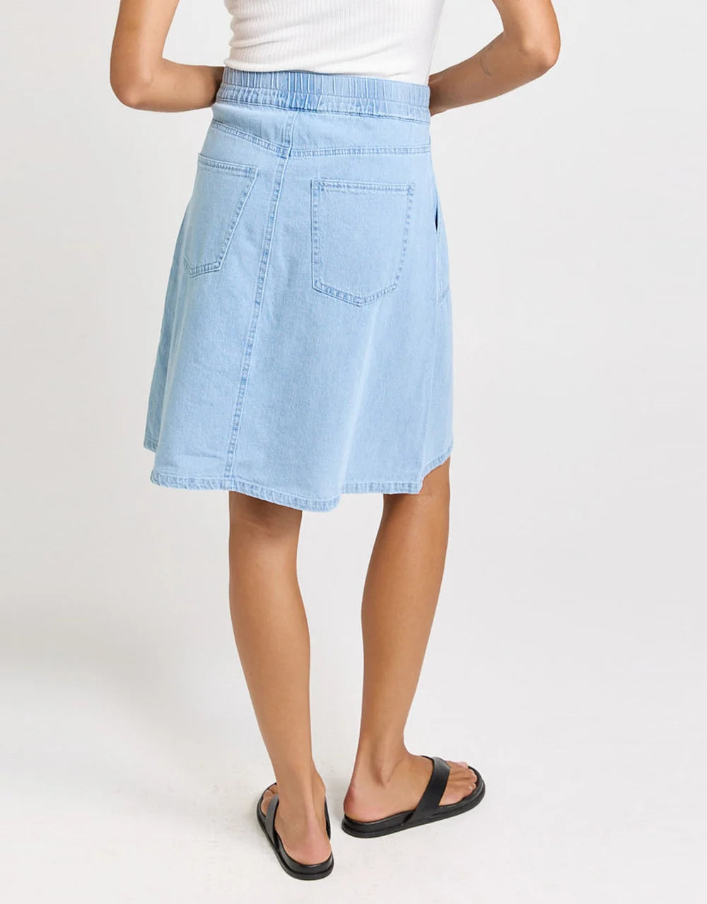 Image of A woman shown from the back wearing a Betty Basics light blue denim A-line skirt with a drawstring waist and back pockets, paired with a white top and black slide sandals, against a plain white studio background.