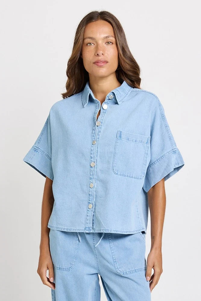 Image of A woman wearing a Betty Basics light blue denim short-sleeve button-up shirt and matching light blue denim pants, photographed against a clean light gray studio background.