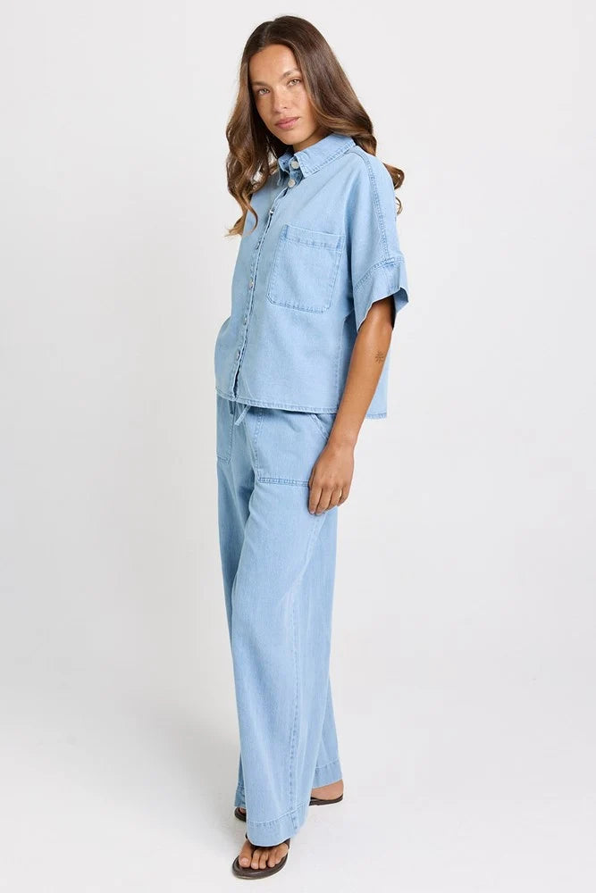 A woman modeling a Betty Basics light blue denim matching set consisting of a short-sleeve button-up shirt and wide-leg pants, photographed against a plain white studio background.