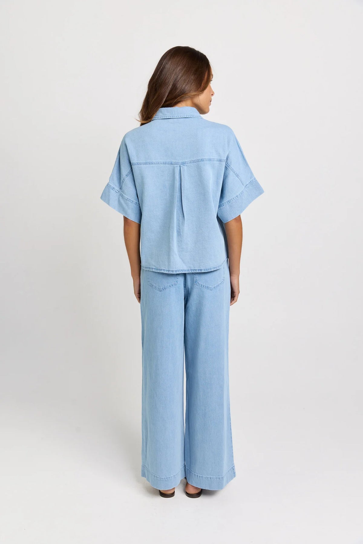 A woman viewed from the back wearing a Betty Basics light blue denim short-sleeve button-up shirt and matching wide-leg pants, photographed against a plain light gray studio background.