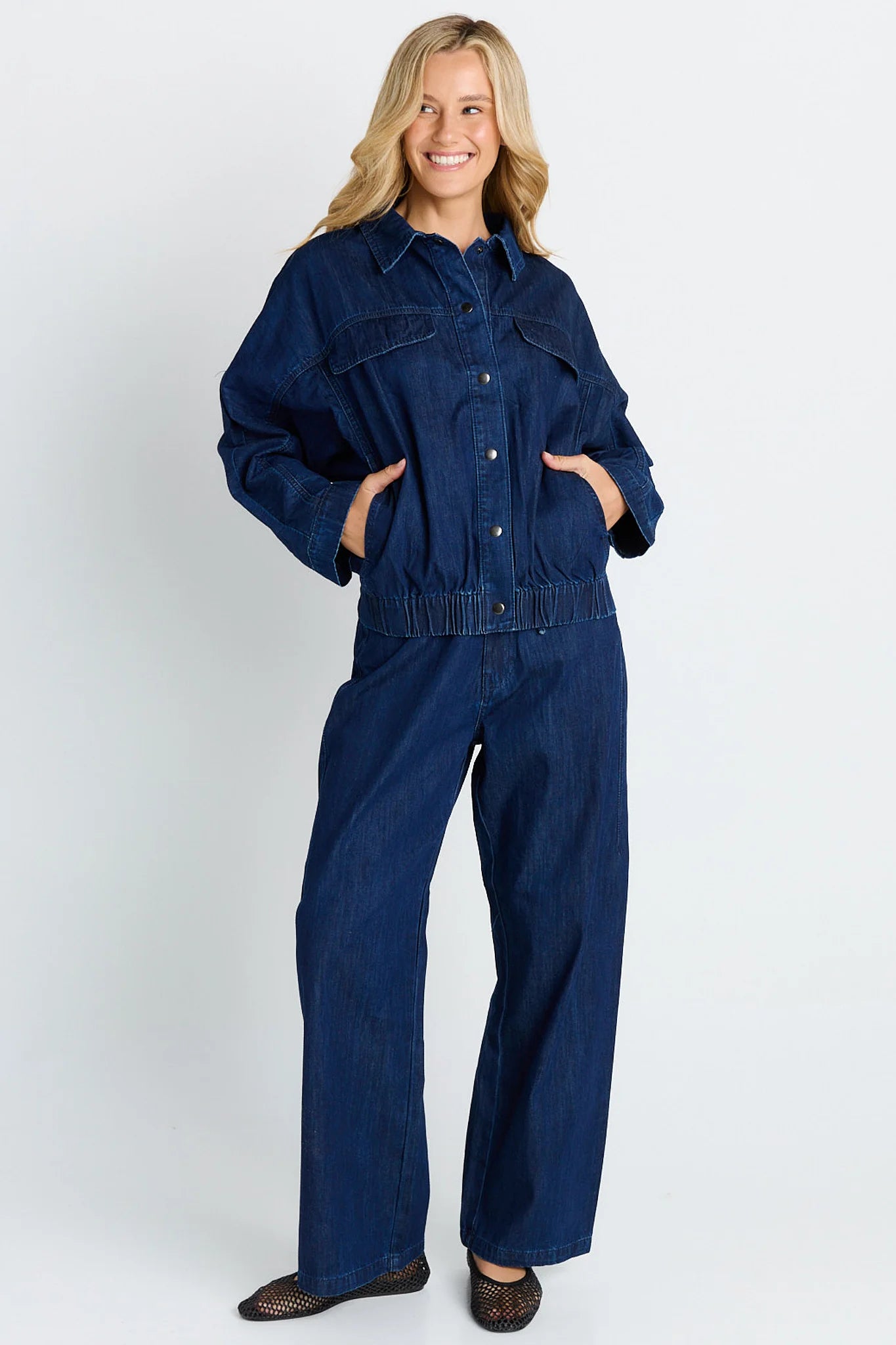 A woman modeling a Betty Basics dark blue denim jeans with a button-up front, chest pockets, and wide legs, standing against a light gray studio background.