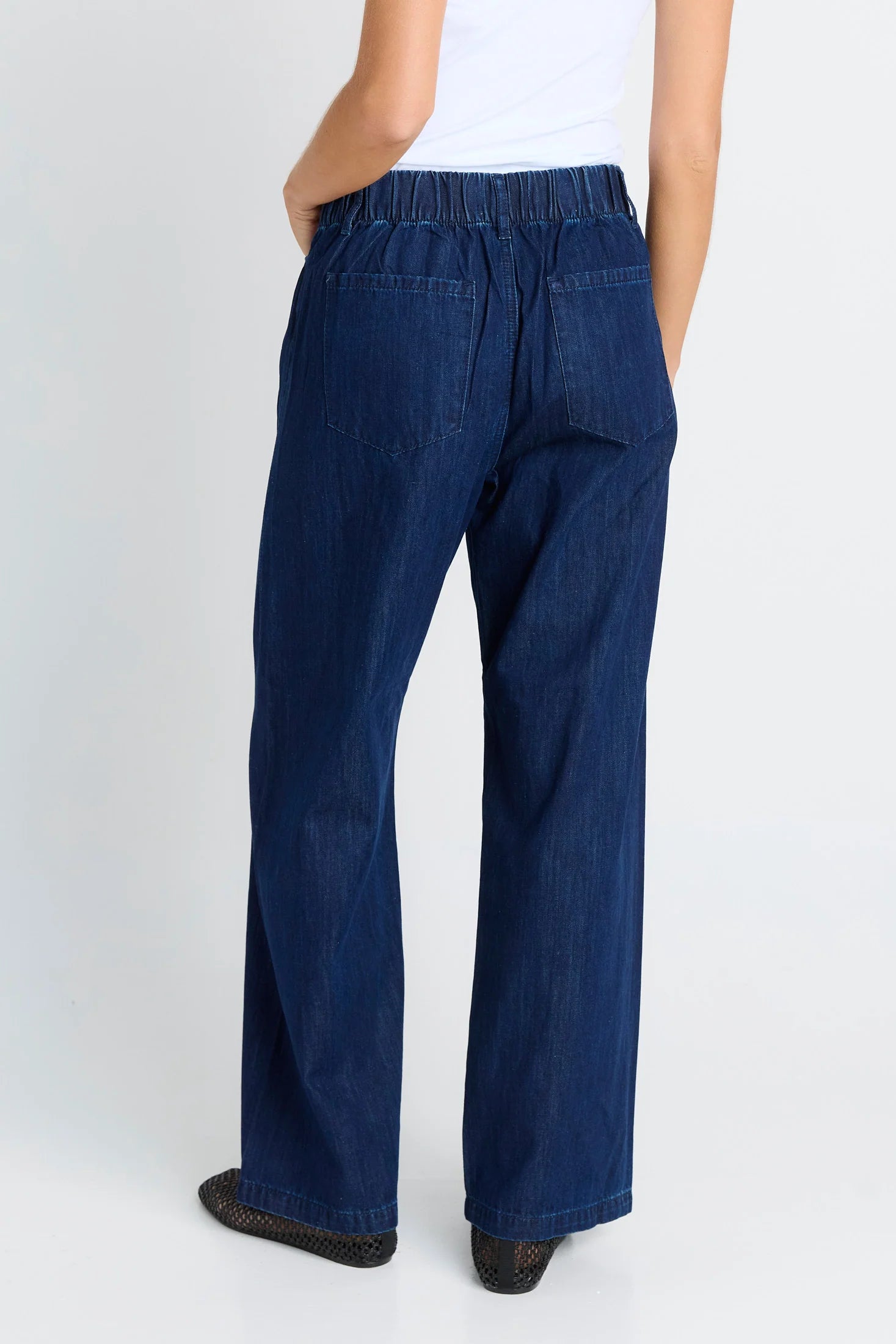 A woman shown from the back wearing Betty Basics dark blue wide-leg denim jeans with an elastic waistband, a white top, and black shoes, against a light gray studio background.