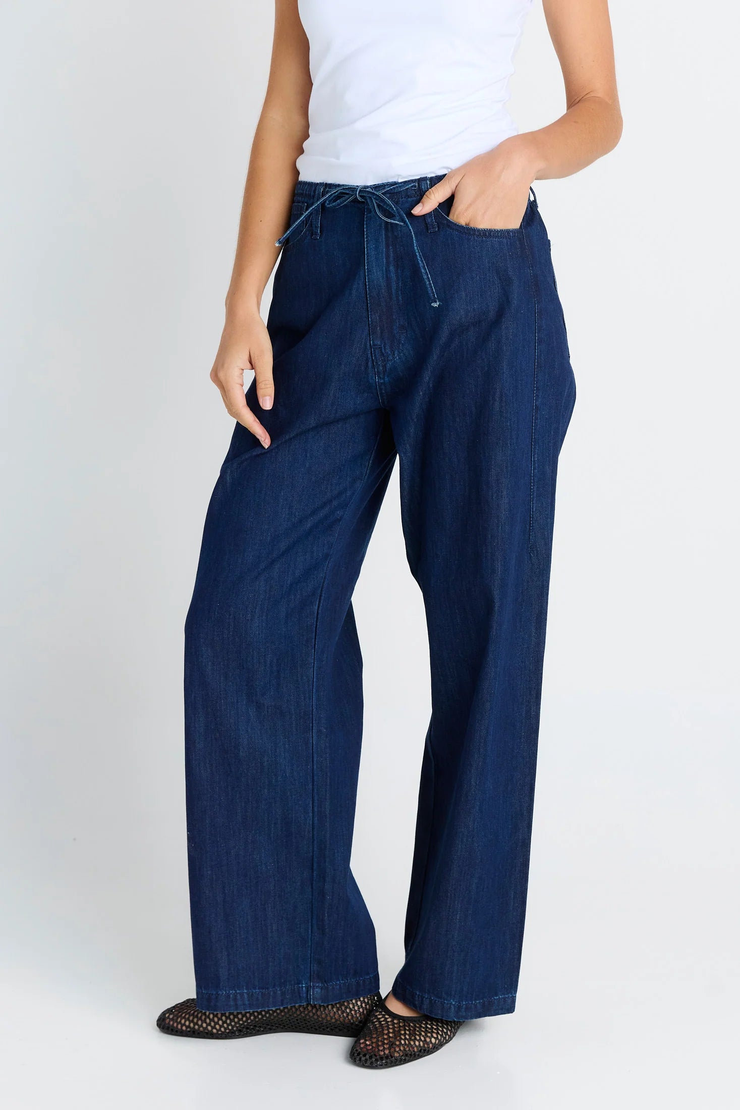 A model wearing Betty Basics dark blue wide-leg denim pants with a drawstring waist, paired with a white tank top and black woven flats, shown from the waist down against a light gray background.