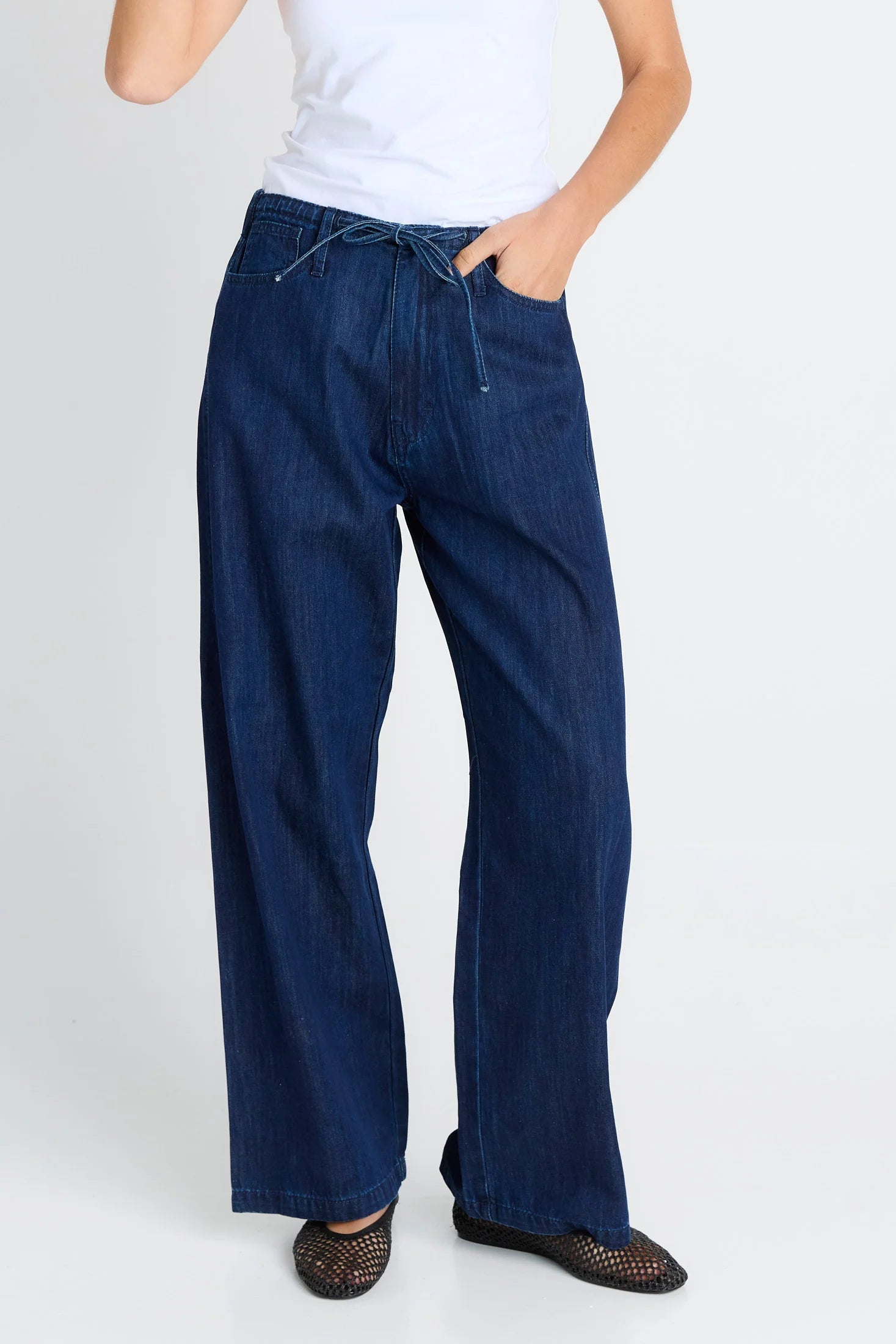 Image of A woman wearing Betty Basics dark blue wide-leg denim jeans with a drawstring waist, paired with a white top and black shoes, shown against a light gray studio background.