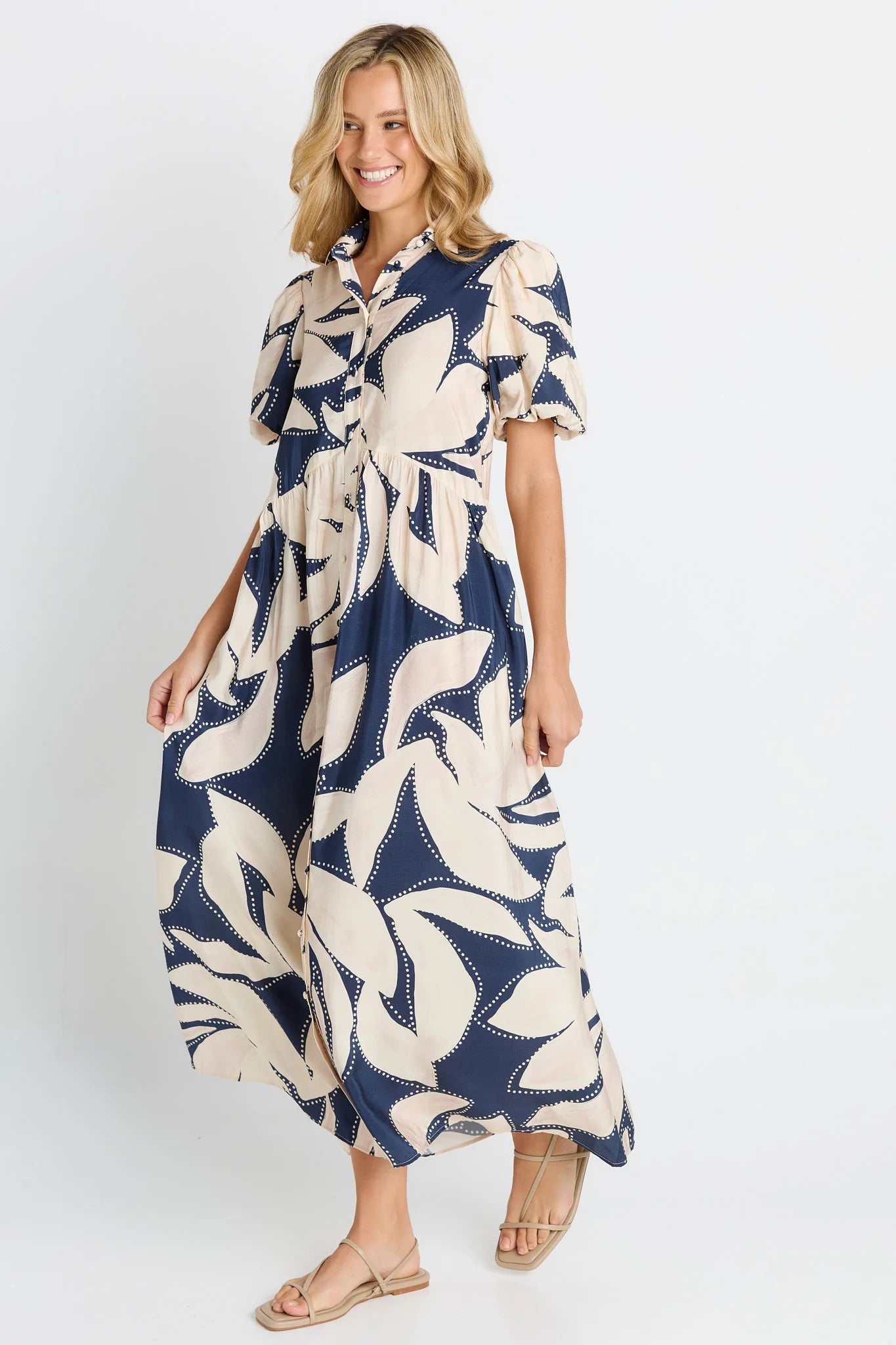 Image of A woman modeling a Betty Basics navy blue and cream leaf-patterned midi dress with short puff sleeves and a button-up collar, paired with beige flat sandals, against a light gray studio background.