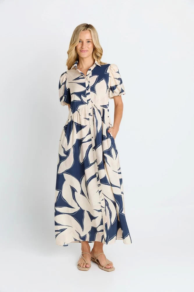 Image of A woman modeling a Betty Basics navy blue and cream tropical leaf print midi dress with short sleeves, a button-up collar, and a tie waist, paired with tan sandals against a plain white studio background.