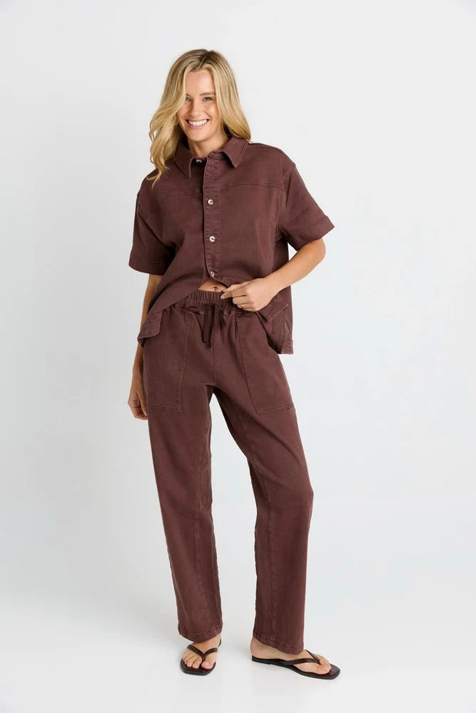 Image of A woman modeling a Betty Basics matching brown short-sleeve button-up shirt and drawstring pants set, paired with black flip-flop sandals, against a plain light gray studio background.