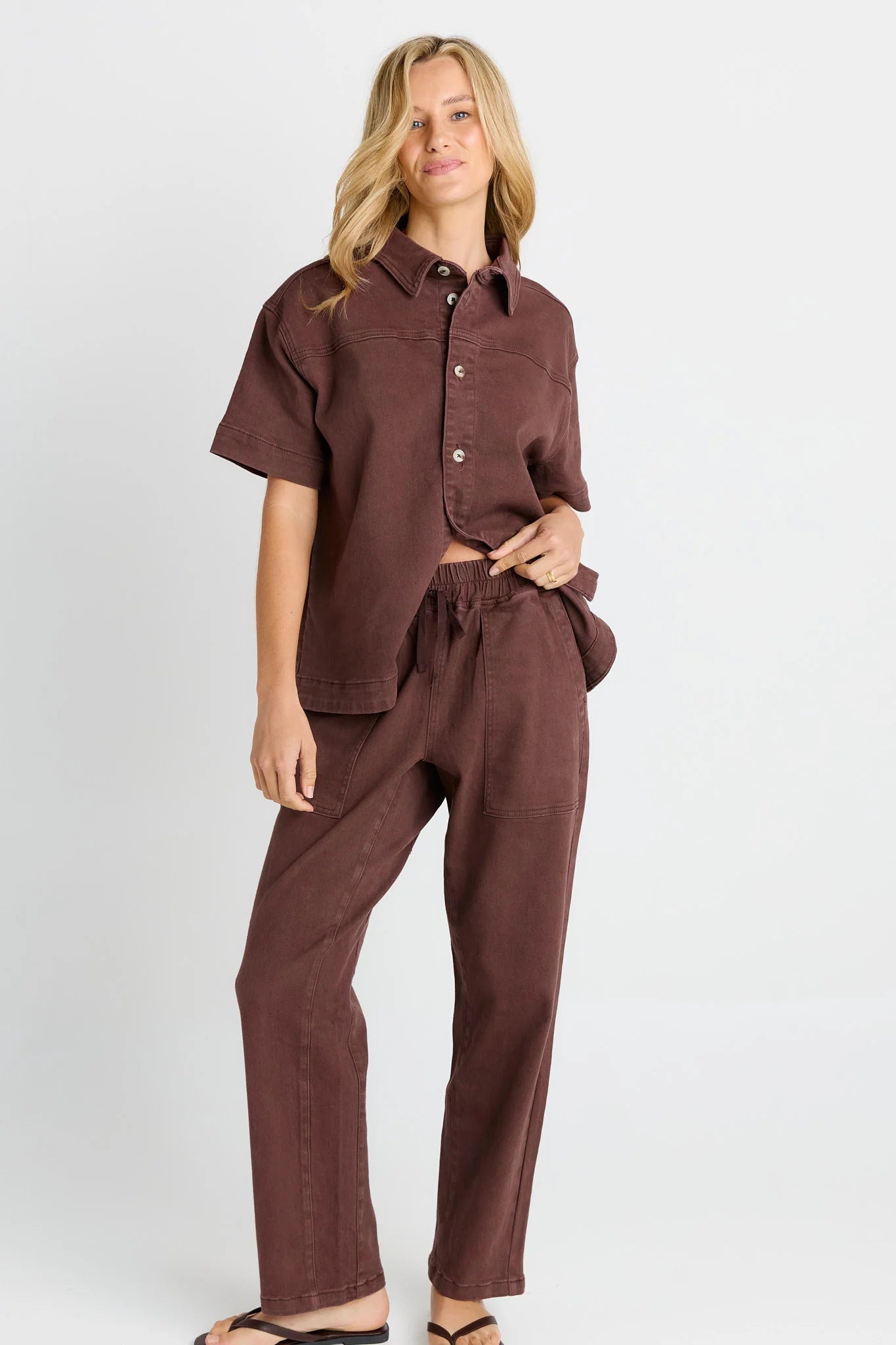 A woman modeling a Betty Basics matching two-piece lounge set in a deep brownish-purple color, featuring a short-sleeve button-up shirt and coordinating drawstring pants, against a light gray studio background.