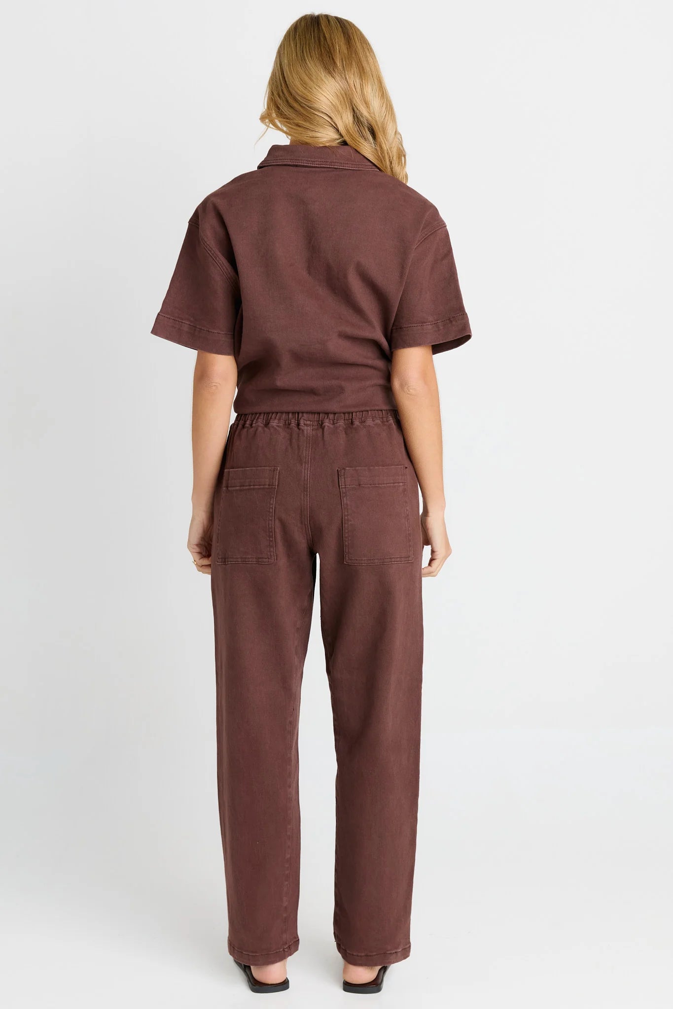A woman viewed from the back wearing a Betty Basics short-sleeve collared top and matching wide-leg pants in a dusty rose-brown color, against a plain light gray studio background.