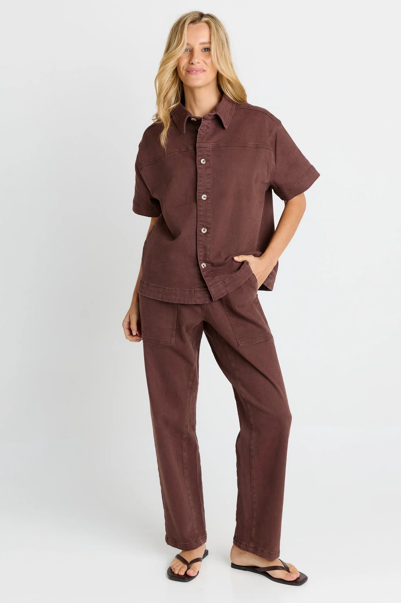 Image of A woman modeling a Betty Basics matching two-piece lounge set in a dusty mauve-brown color, featuring a short-sleeve button-up shirt and wide-leg pants, paired with black flip-flops against a light gray studio background.