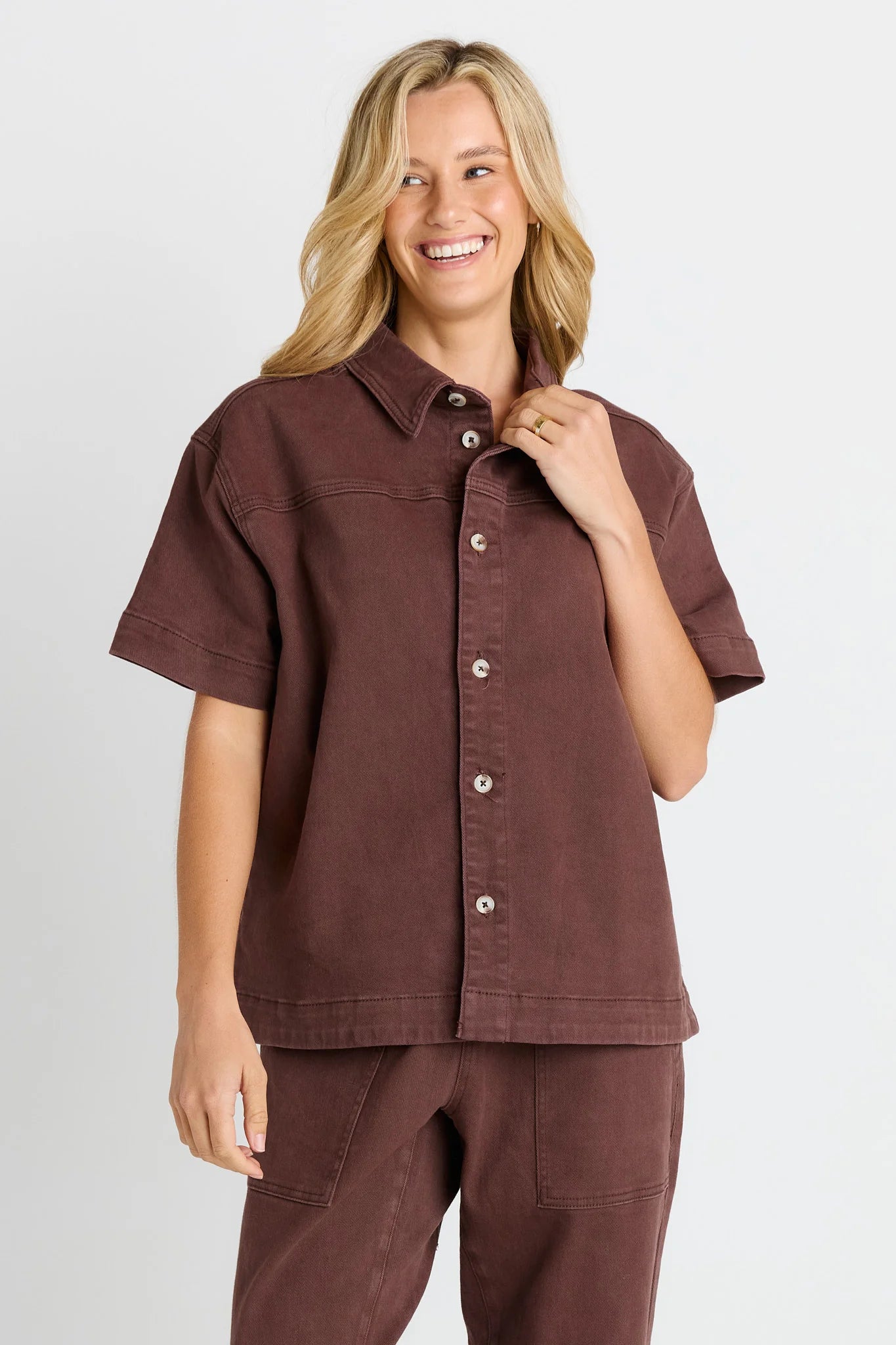 A woman wearing a Betty Basics short-sleeve button-front shirt and matching pants in a dark brown color, photographed against a light gray studio background.