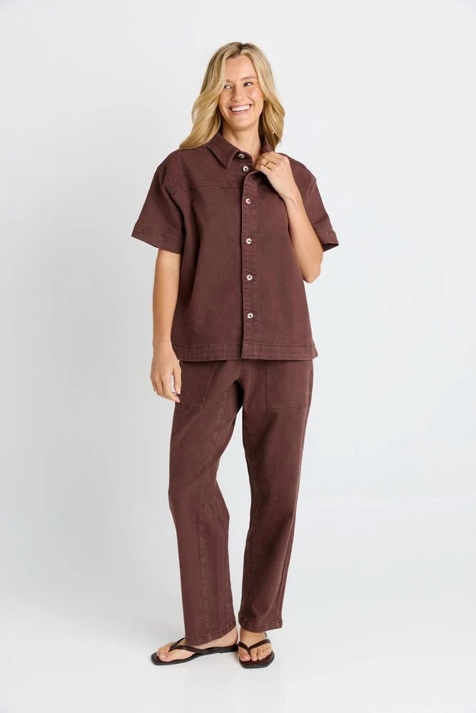 Image of A woman modeling a Betty Basics matching short-sleeve button-up shirt and wide-leg pants set in a dark brown color, standing against a light gray studio background.