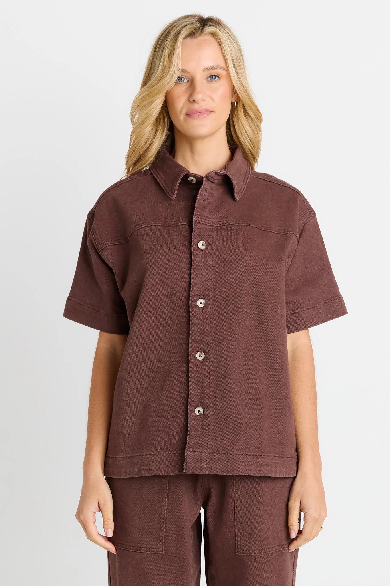 Image of A woman modeling a Betty Basics short-sleeve button-up shirt in a muted dusty rose-brown color, paired with matching pants, against a light gray studio background.