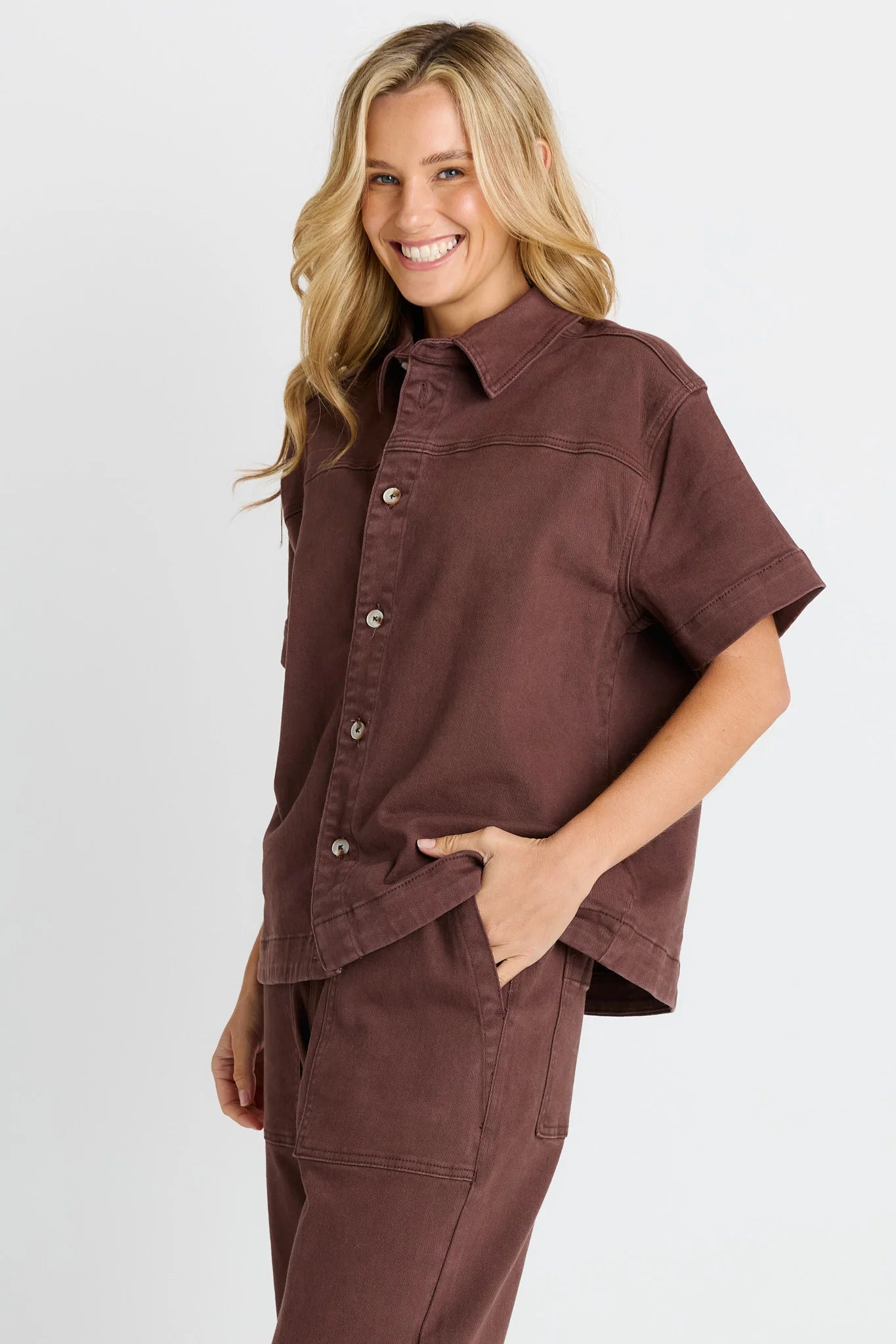Image of A woman modeling a Betty Basics short-sleeve button-front shirt and matching pants in a dusty rose-brown color, photographed against a clean light gray studio background.