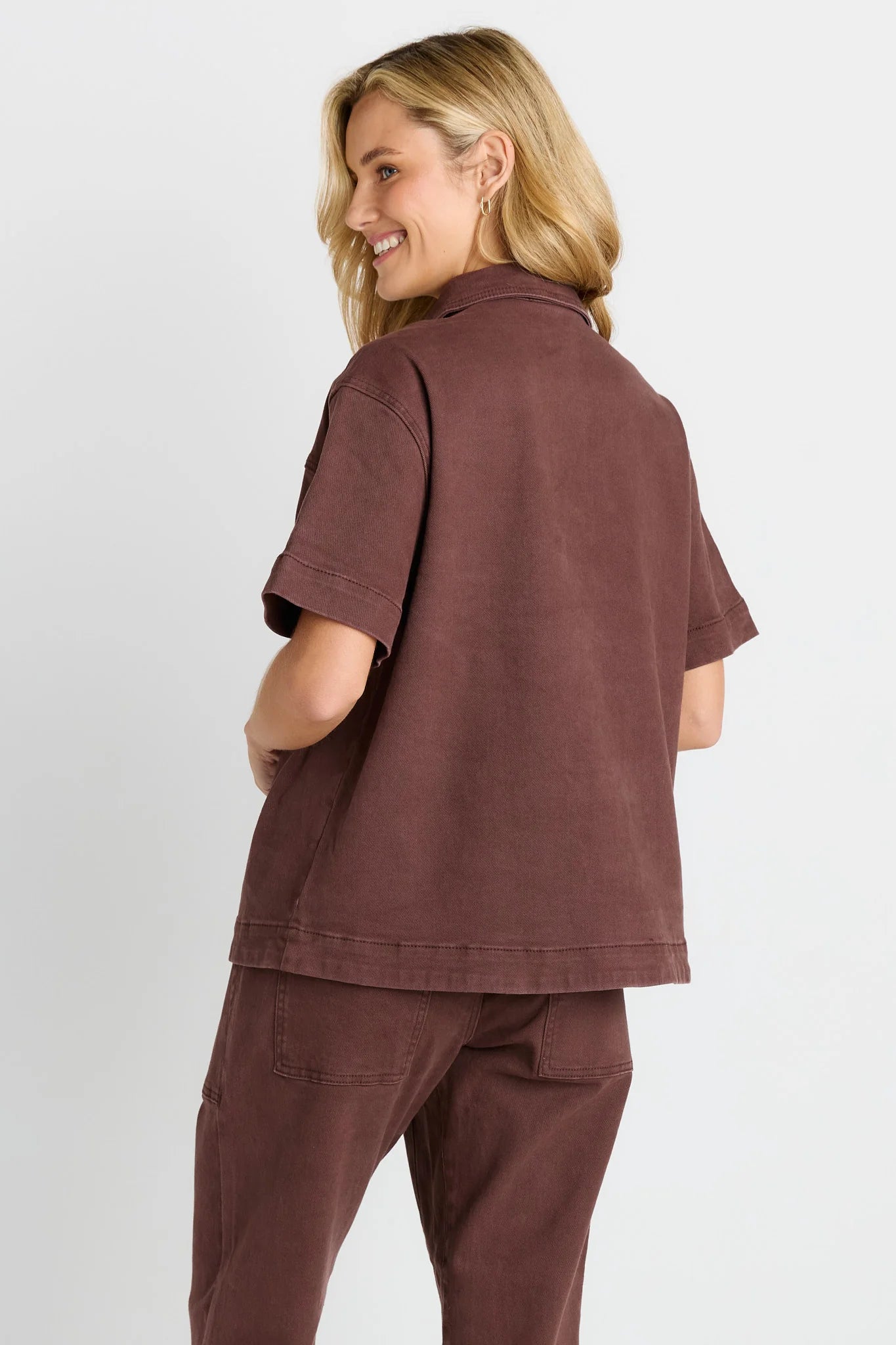 Image of A woman wearing a Betty Basics 
matching rust-brown short-sleeve button-up shirt and pants, shown from a back-three-quarter angle against a light gray studio background.
