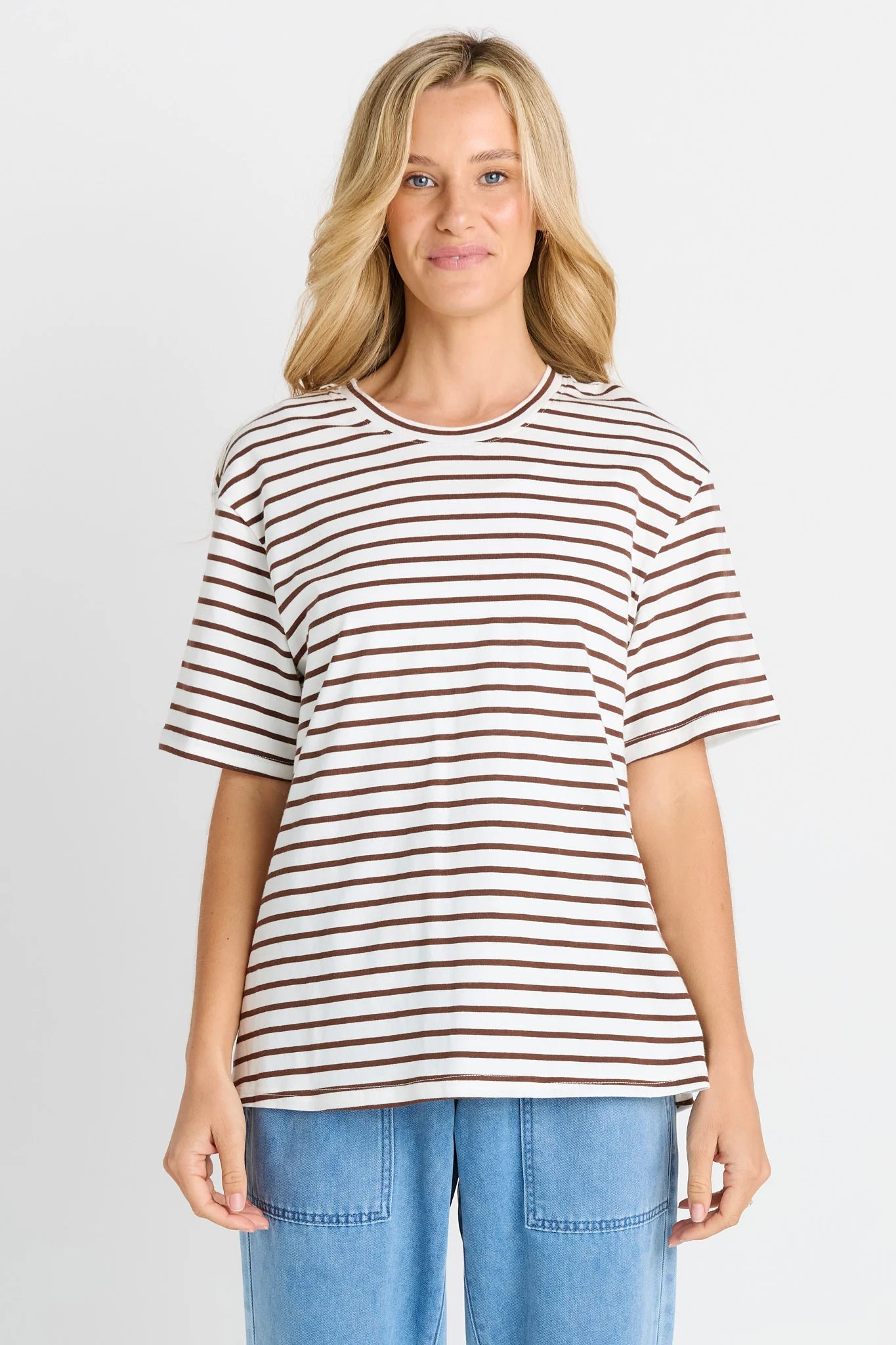 Image of A woman modeling a Betty Basic short-sleeve crewneck t-shirt with thin brown horizontal stripes on a white background, paired with light blue jeans, against a light gray studio backdrop.