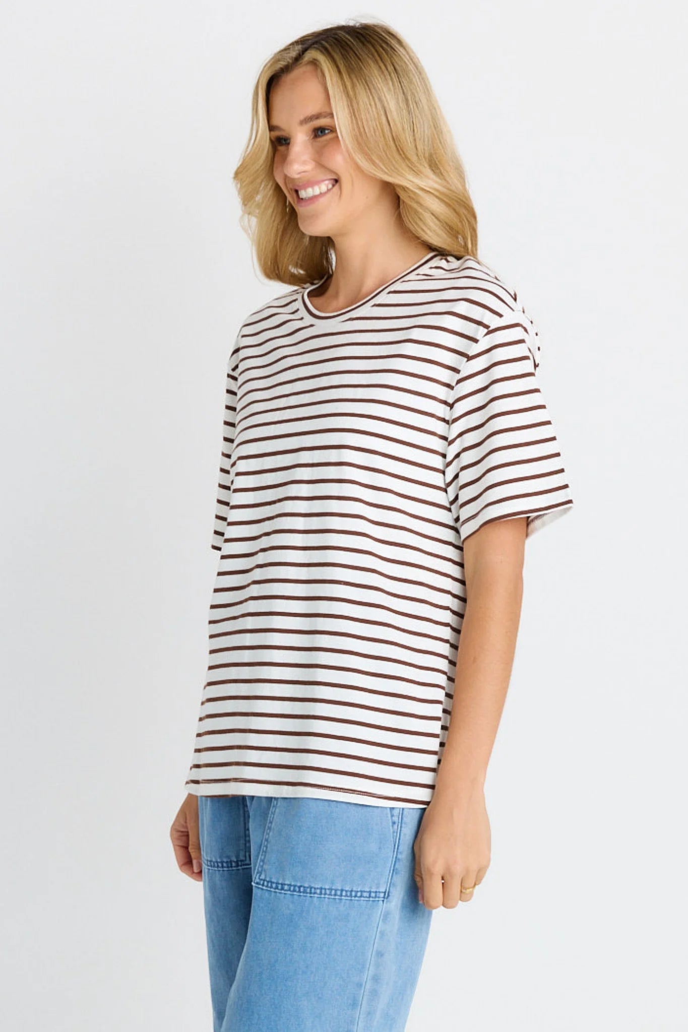 Image of A woman wearing a Betty Basics short-sleeve crewneck t-shirt with thin brown horizontal stripes on an off-white base, paired with light blue jeans, photographed against a light gray studio background.