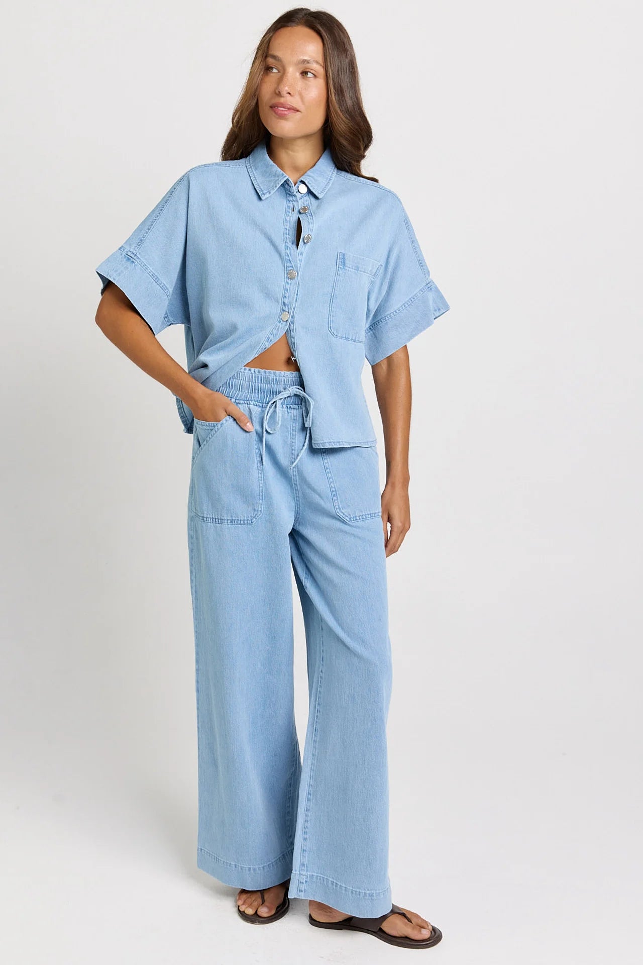 Image of A woman modeling a Betty Basics light blue denim two-piece set consisting of a short-sleeve button-up shirt and matching wide-leg drawstring pants, photographed against a plain light gray studio background.
